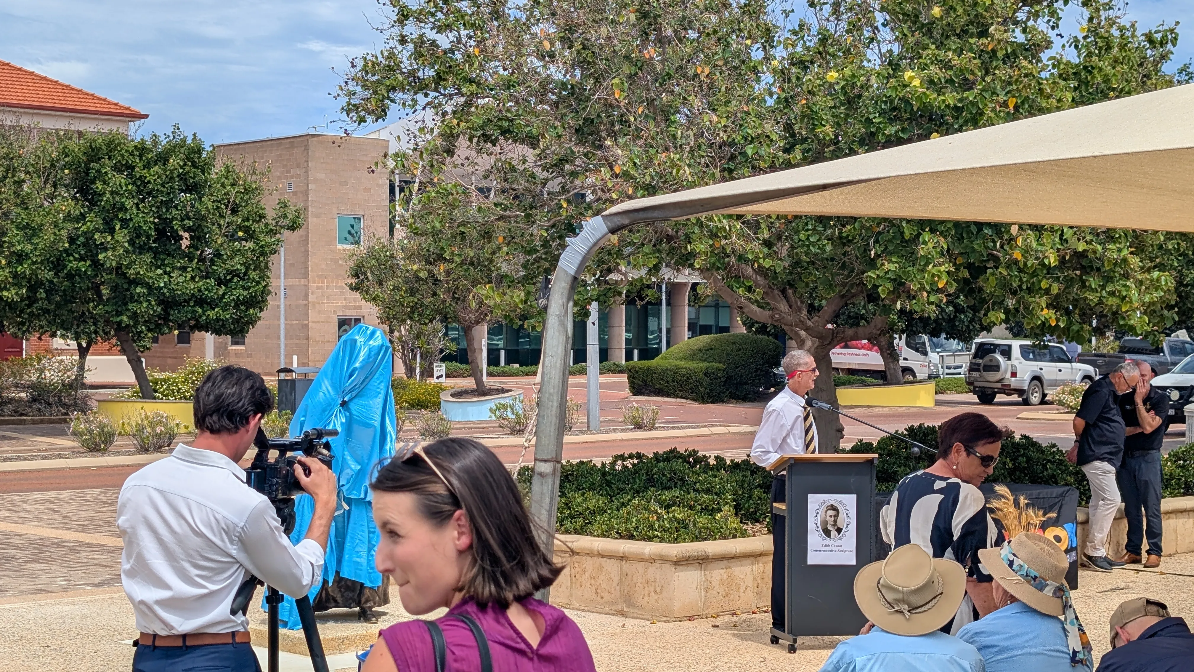 A photo of a statue covered by a blue tarpaulin surrounded by people at an unveiling ceremony.