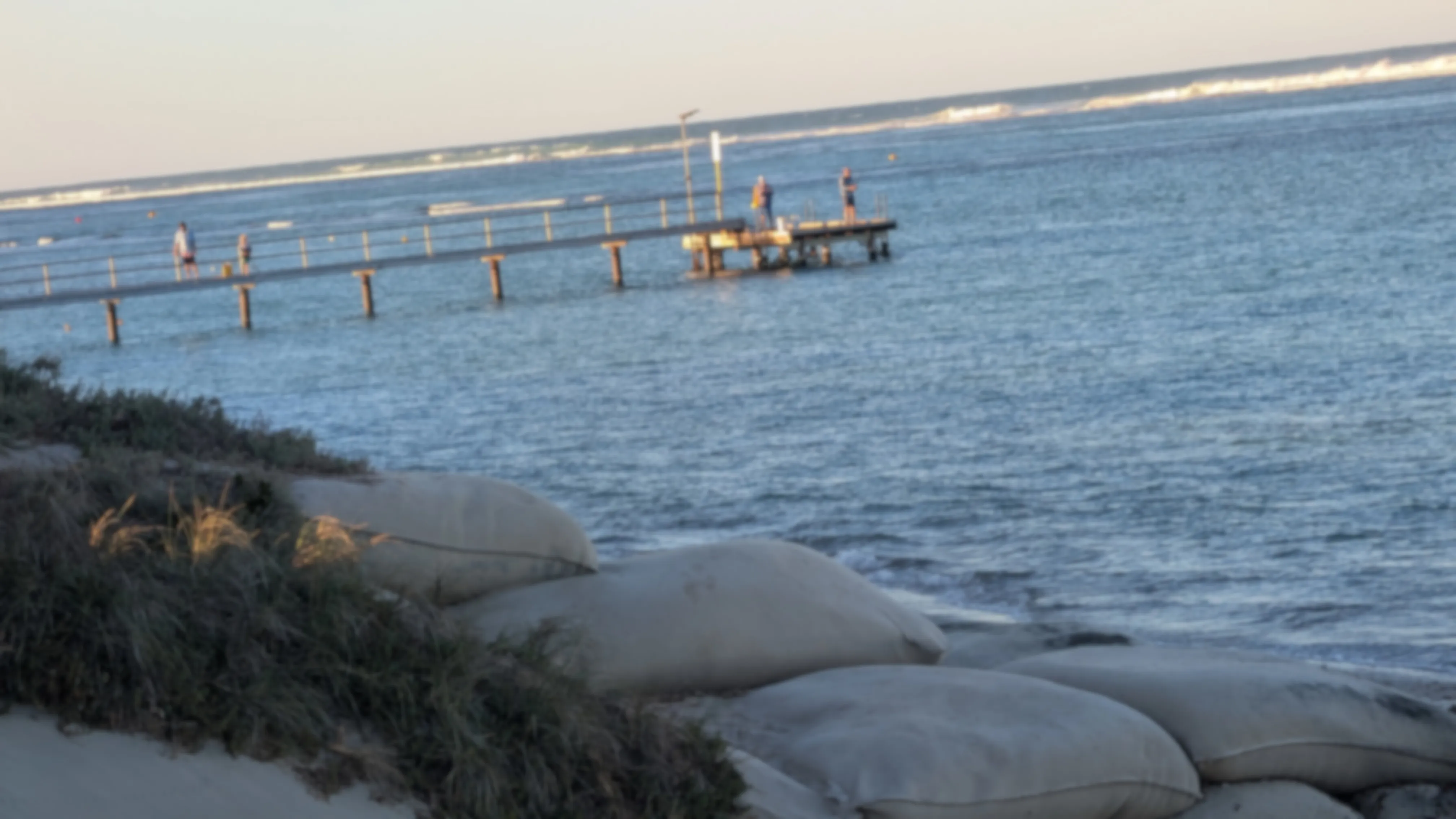 A blurred photo of a jetty with four people fishing off of it into the ocean.